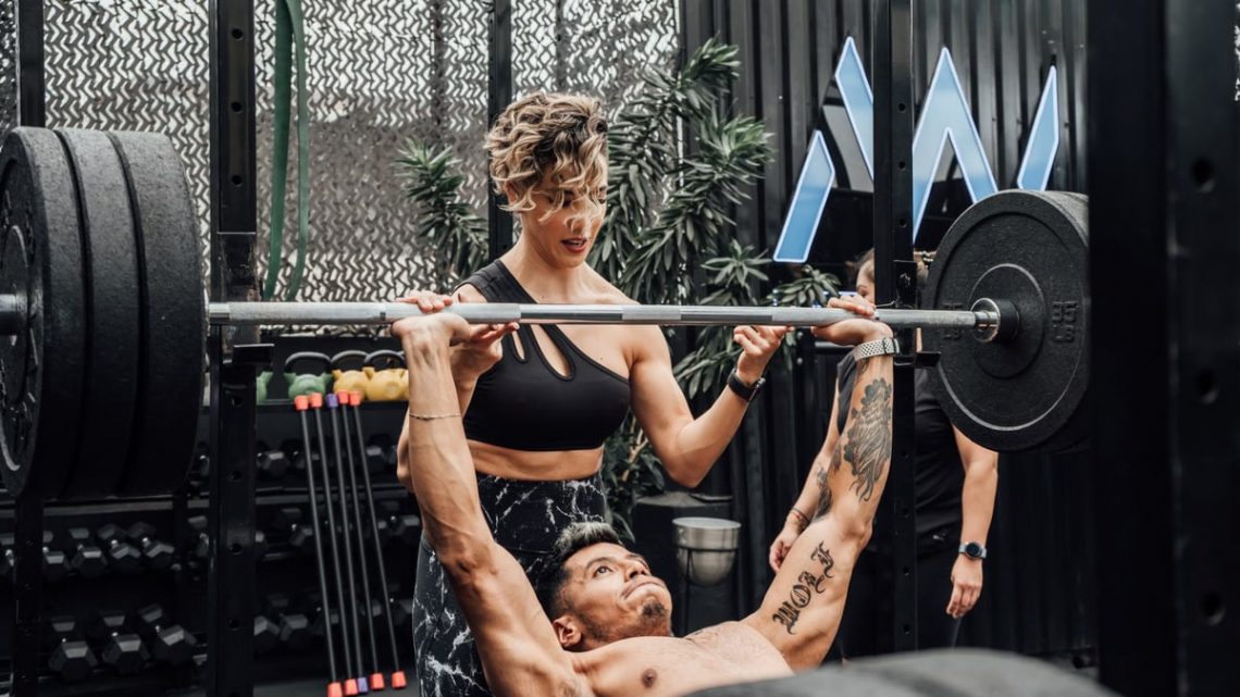 Man lifting weights with woman spotting in Mexico City gym. Urban and dynamic fitness scene.